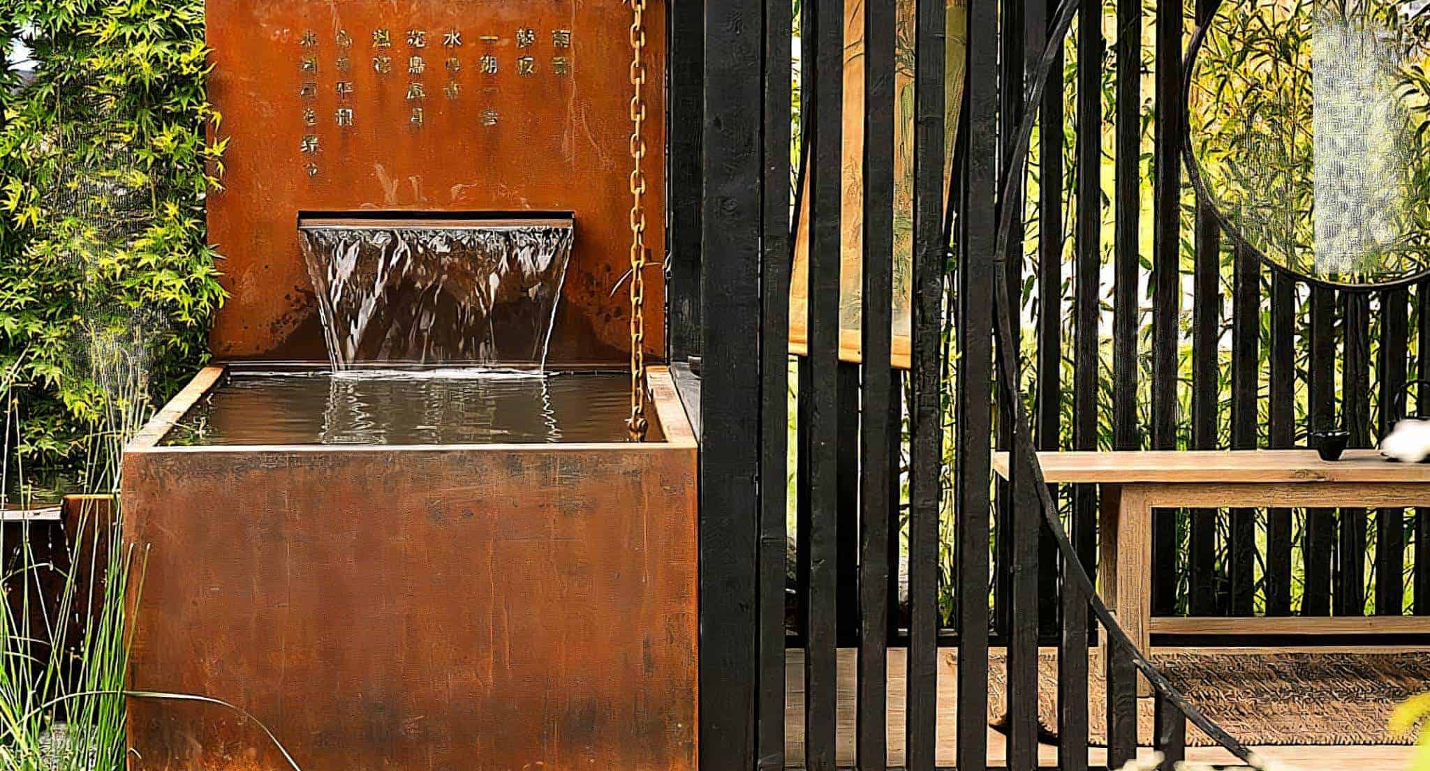 Rustic metal water feature with Japanese inscriptions, water cascading into a rectangular basin, framed by tall black wooden slats and lush greenery.